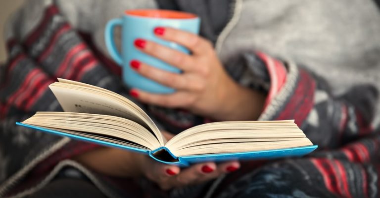 a woman wrapped in a blanket is holding a book open and holding a mug in her other hand