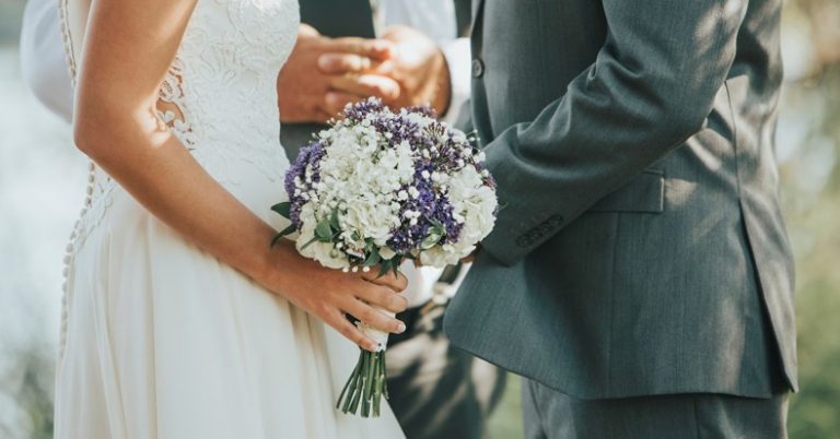 A couple in the process of getting married in front of an ordained minister is facing each other with the woman holding a bouquet