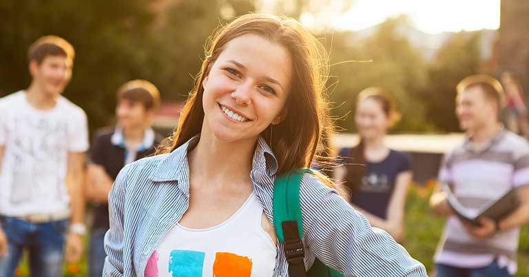 female with a backpack on one shoulder stands in foreground as fellow peers her age stand in the background conversating