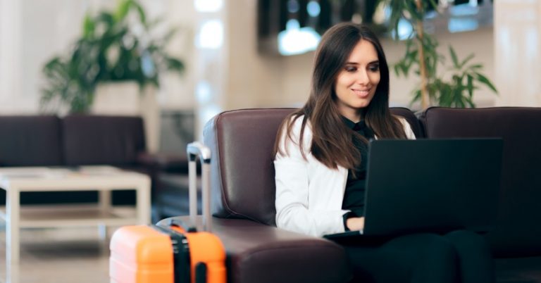 a woman with a laptop in her lap is sitting on a couch with her rolling suitcase next to her