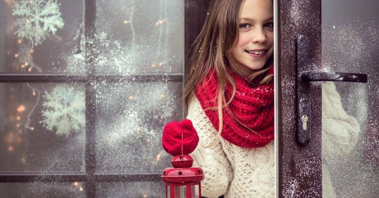 A girl wearing festive clothing peeks from a winter and is holding a lantern