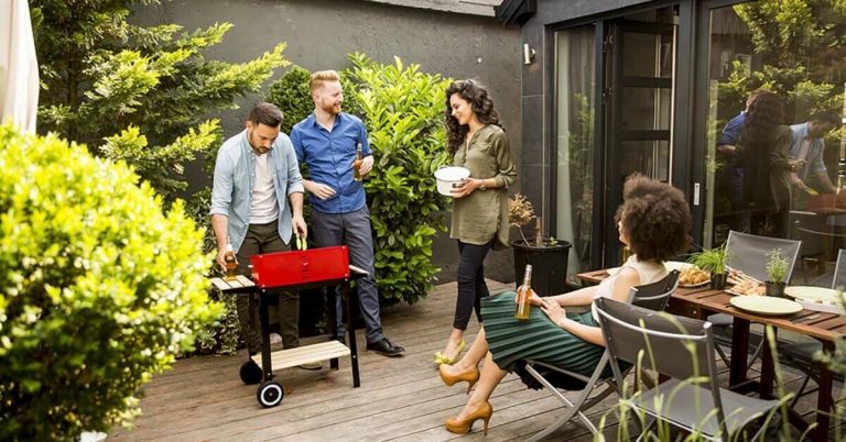 A group of young adults stand around a grill chatting.