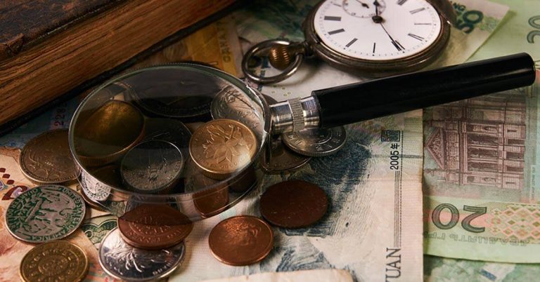 A collection of coins and foreign paper money sit on a table next to a magnifying glass.