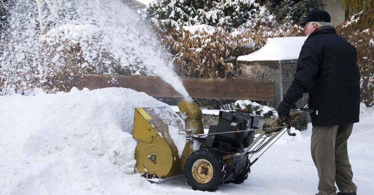 An older man uses a snow blower to clear his driveway.