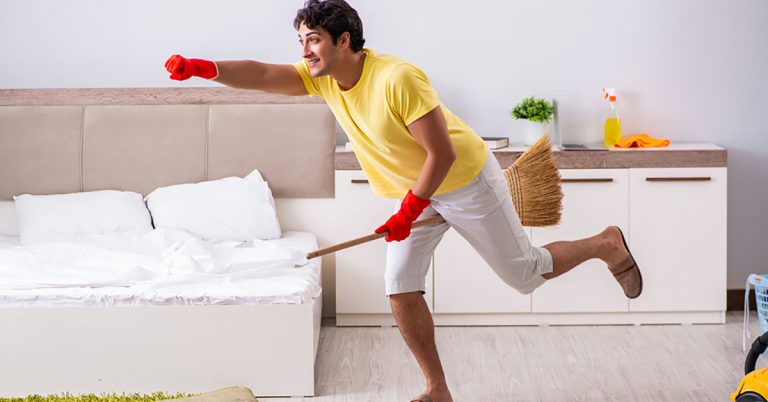 A young man pretends to ride a broom while cleaning his bedroom.