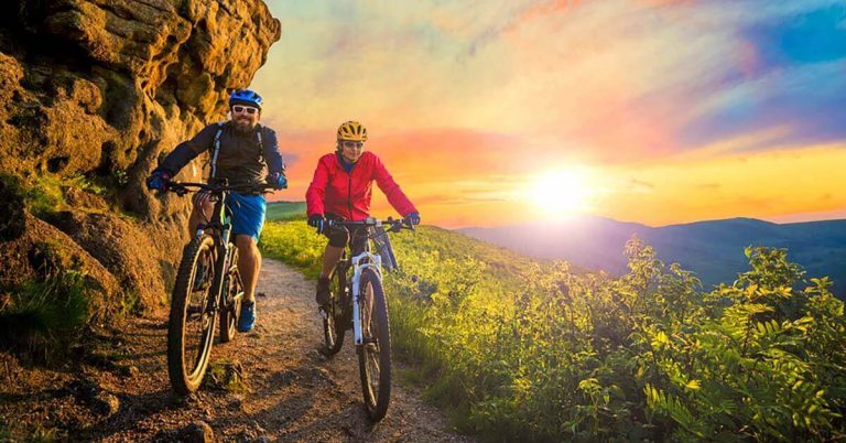 An older couple goes mountain biking as the sun sets behind them.