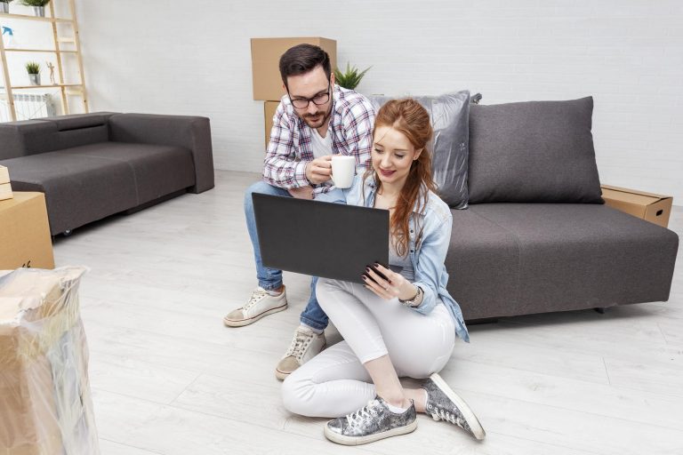 a couple sitting on on the floor and on the couch surrounded by boxes are both looking at a computer screen while they weigh their storage options