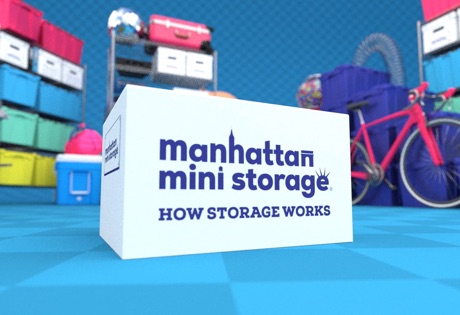 A cardboard moving box reading "Manhattan Mini Storage, how storage works" with shelves of boxes in the background