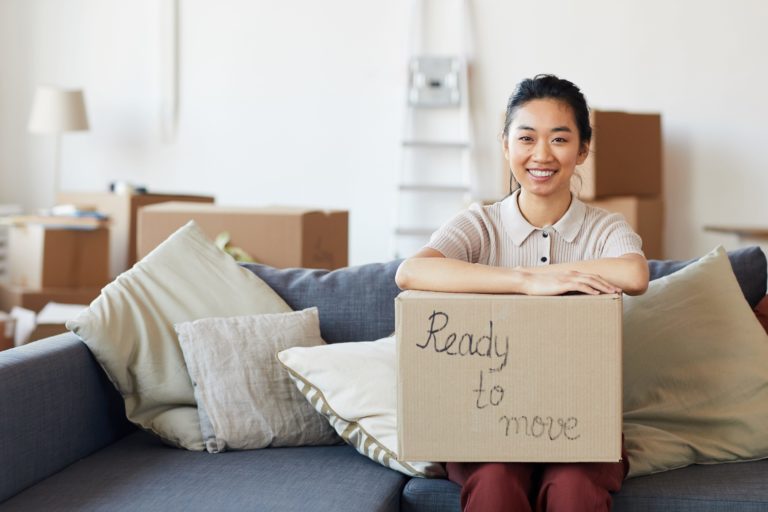 Young female sits on couch with box labeled "Ready to Move" wiht more packed boxes in the background