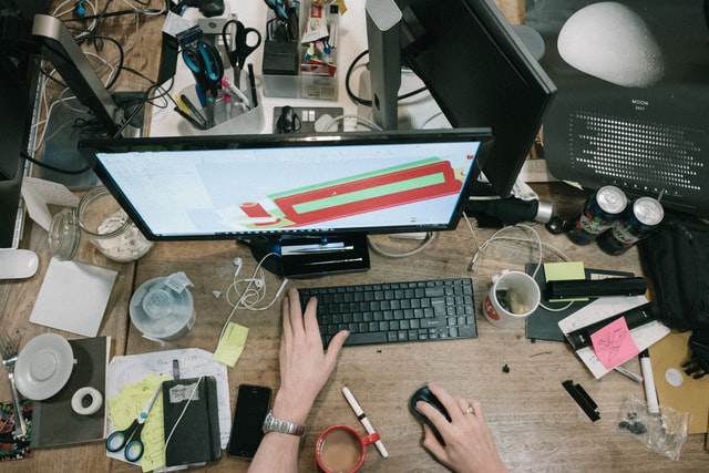 Person working at a messy desk surrounded by clutter.