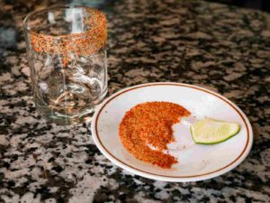 A plate with a lime wedge and a mix of raw sugar and Tajin rests next to a rocks glass with half of the cup rip covered in the mixture.