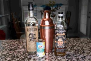 A frosty copper shaker stands upright with a bottle of Cimarron Tequila, Mathidle Orange Liqueur, and a can of Dole Pineapple juice. 