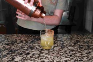 A woman pours her cocktail out of her shaker and into a prepared rocks glass. 