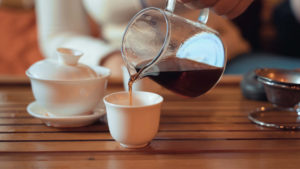 A white saucer with a cup and lid brews tea, while the small glass pitcher pours perfectly made tea into a tasting cup. 